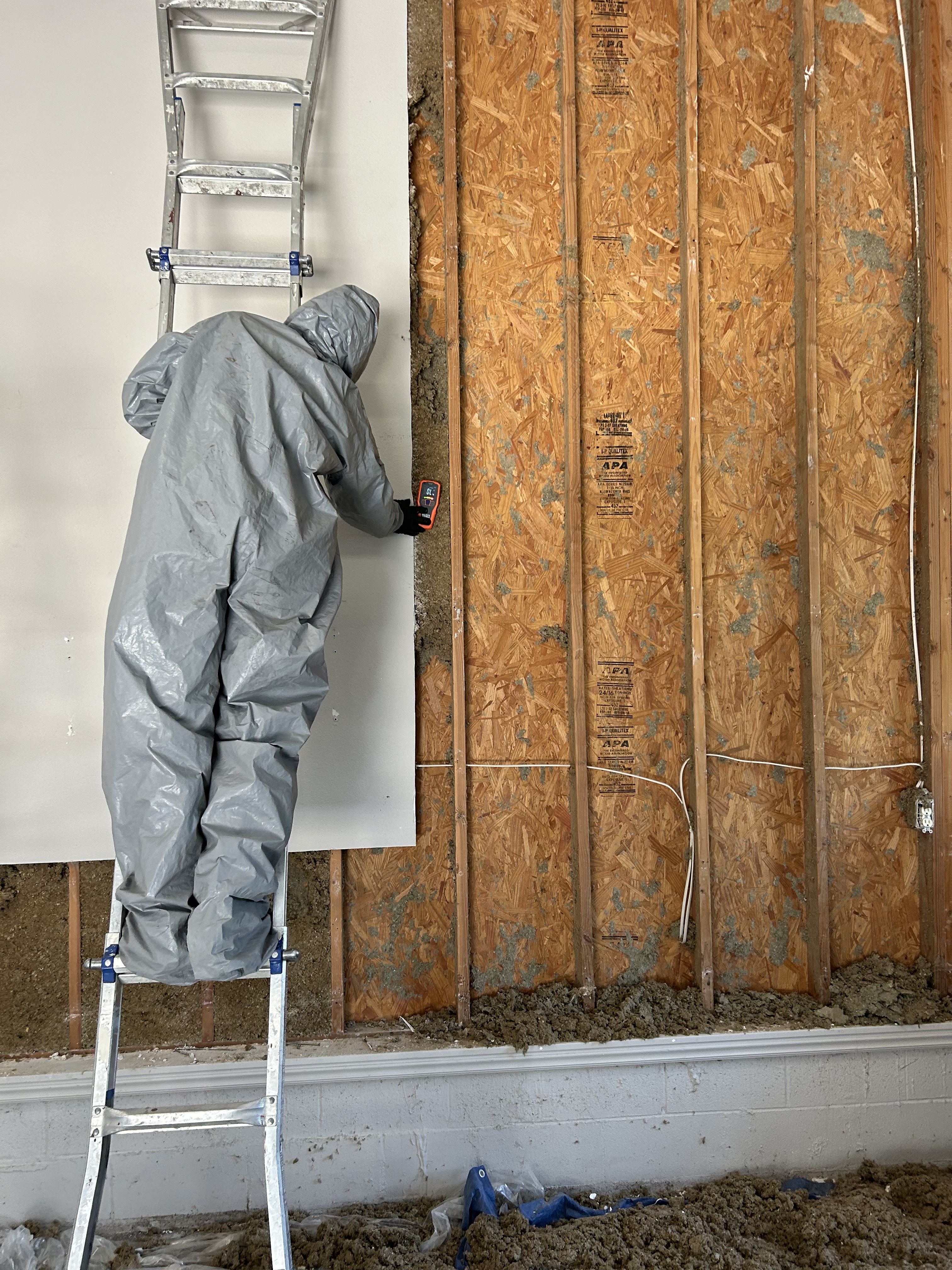 A worker in a gray jumpsuit stands on a ladder, inspecting or repairing a wall with padding behind the drywall. A level is...